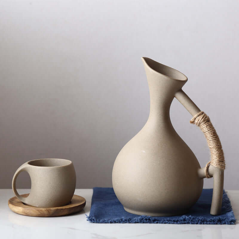 Handcrafted ceramic kettle and cup set in minimalist Nordic style on blue cloth, perfect for serving cold beverages.