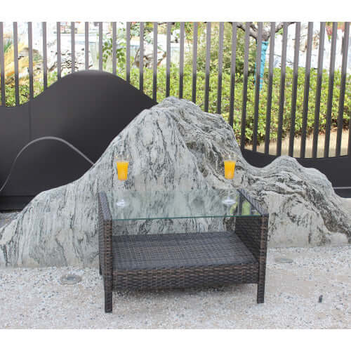 35 inch rectangular coffee table made of weather-resistant brown PE rattan with a tempered glass top, set against a stone backdrop.